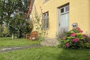 Un bâtiment jaune avec une fenêtre, une chaise en bois et un jardin avec des fleurs roses.