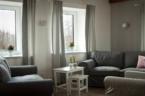 Ein Wohnzimmer mit einem grauen Sofa, einem weißen Tisch mit Blumen und einem Fenster mit grauen Vorhängen.