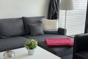Ein graues Sofa mit einem weißen Kissen mit dem Aufdruck 'Strandgut', einer roten Decke, einem weißen Tisch mit einem kugelförmigen Gegenstand und einer kleinen Topfpflanze.