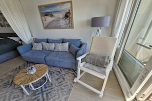 A living room with a blue sofa, a round coffee table, a beige armchair, a lamp, and a painting of a beach scene. - South Beach happiness (Burgtiefe-Südstrand)