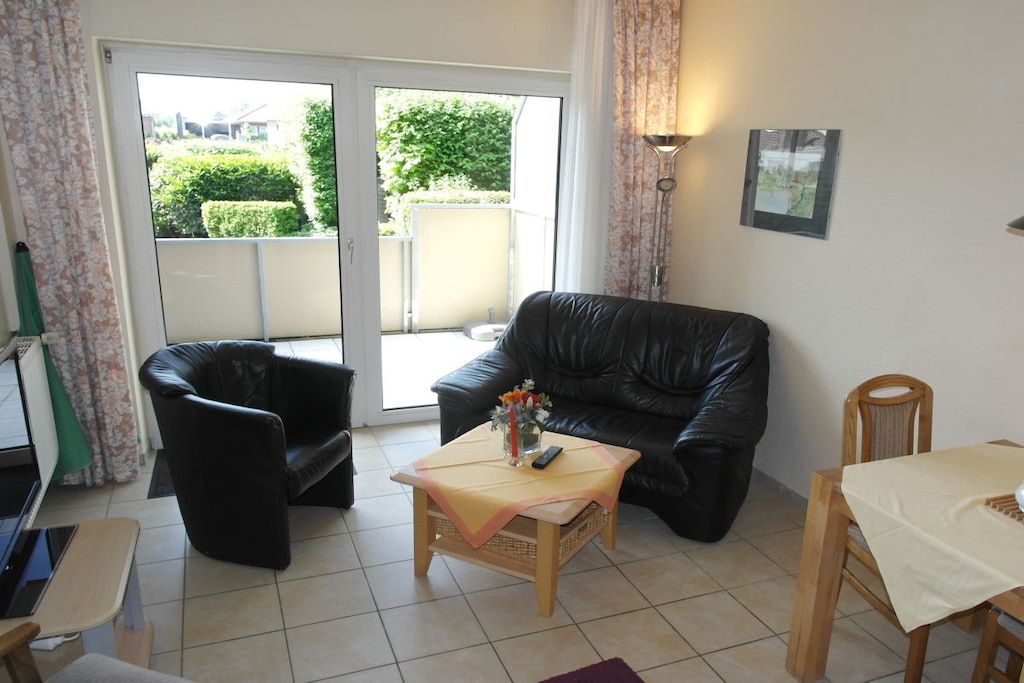 A living room with a black leather sofa, a small wooden table with a vase of flowers, a dining table with a tablecloth, and a sliding glass door leading to a balcony.