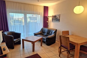 Ein Wohnzimmer mit Sofa, Couchtisch, Esstisch mit Stühlen und einem großen Fenster mit Vorhängen.