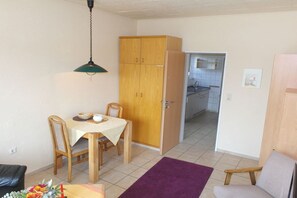 Ein kleines Zimmer mit Esstisch, Stühlen, einem violetten Teppich, einem Holzschrank und einem durch eine offene Tür einsehbarer Küchenbereich.
