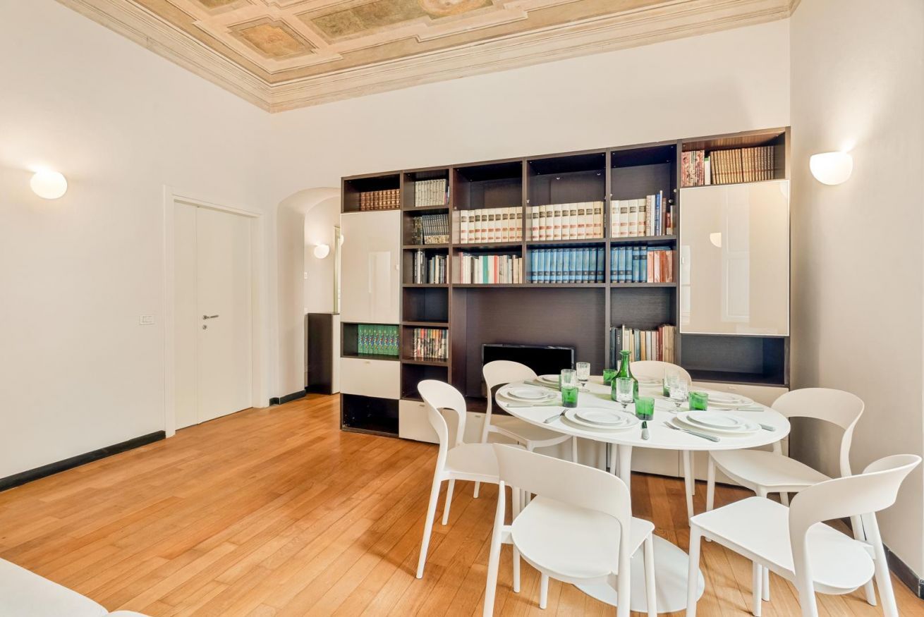 A modern dining area with a round table set for four, surrounded by white chairs, with a bookshelf and a fireplace in the background.