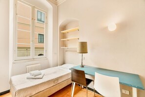 Ein Schlafzimmer mit einem Einzelbett, einem Schreibtisch, einem Stuhl, einer Lampe und einem Fenster mit Blick auf Gebäude.