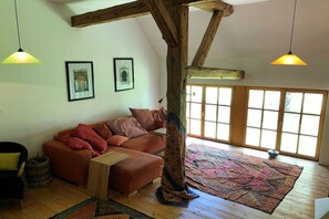 Ein gemütliches Wohnzimmer mit Holzbalkendecke, einer Eckcouch, einem Couchtisch und einem großen Fenster mit Tageslicht.