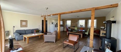 Een ruime woonkamer met een houten plafond, een bank, een salontafel, een open haard en een keuken op de achtergrond.