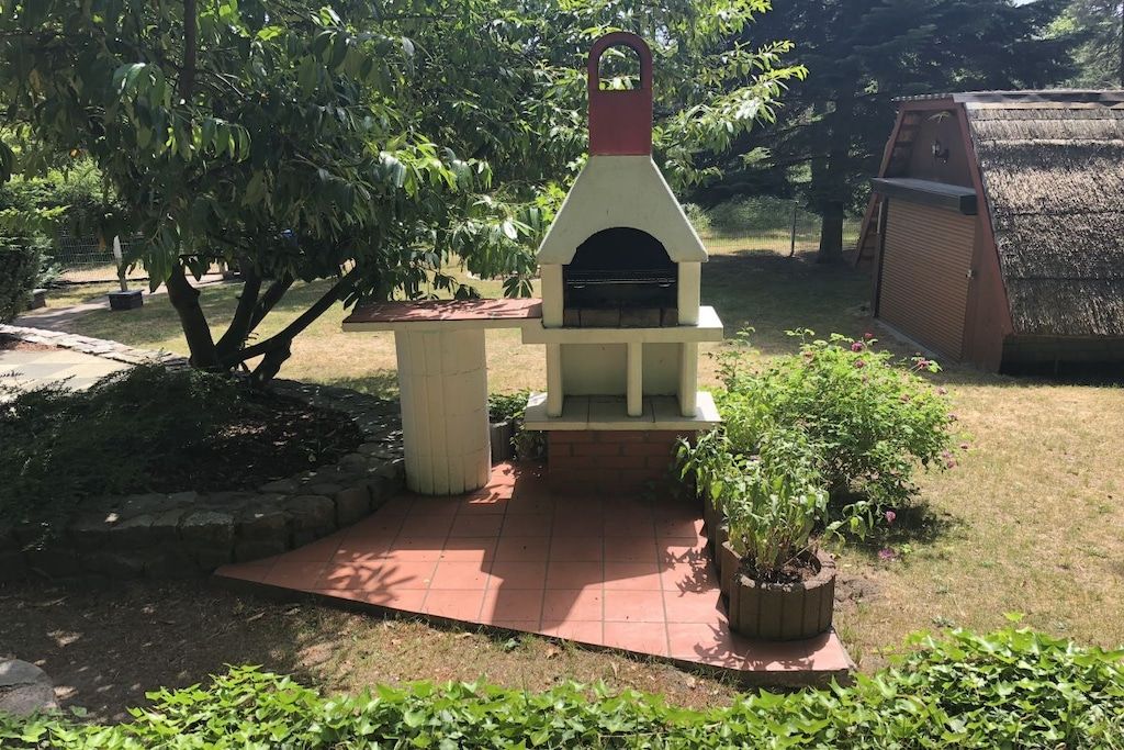 Een bakstenen buitenbarbecue met een witte en rode schoorsteen, omringd door een tuin met een bloempot en een houten constructie op de achtergrond.