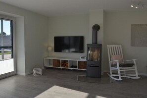 A modern living room with a fireplace, a flat-screen TV mounted on the wall, a white rocking chair, and a window providing natural light. - Holiday apartment with 2 bedrooms (Kronsgaard)