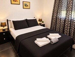 A neatly made bed with a black and white bedspread, two pillows, and two folded towels. There are two bedside tables with lamps, a small potted plant, and two framed pictures on the wall. - Tania's House - Meteora Peaceful Garden Retreat (Flamouli)