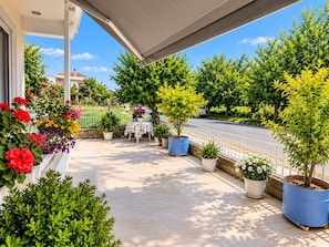 A covered outdoor area with potted plants and flowers, a table with a tablecloth, and a view of a road and greenery in the background. - Tania's House - Meteora Peaceful Garden Retreat (Flamouli)