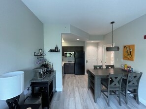 A modern kitchen with dark cabinetry, a central island, and a dining area with a table and chairs. - King & Queen Beds! FREE SHUTTLE * FREE PARKING * No Resort Fee * Coffee Bar * Waffle Bar * 76" & 70" Smart TV's * Pool & Hot Tub * Plush Towels & Linens * Gas BBQ Grill * King Bed Has Adjustable Base * Sleeper Sofa * 2 Mile to Disney Springs *7 Mile (Orlando)