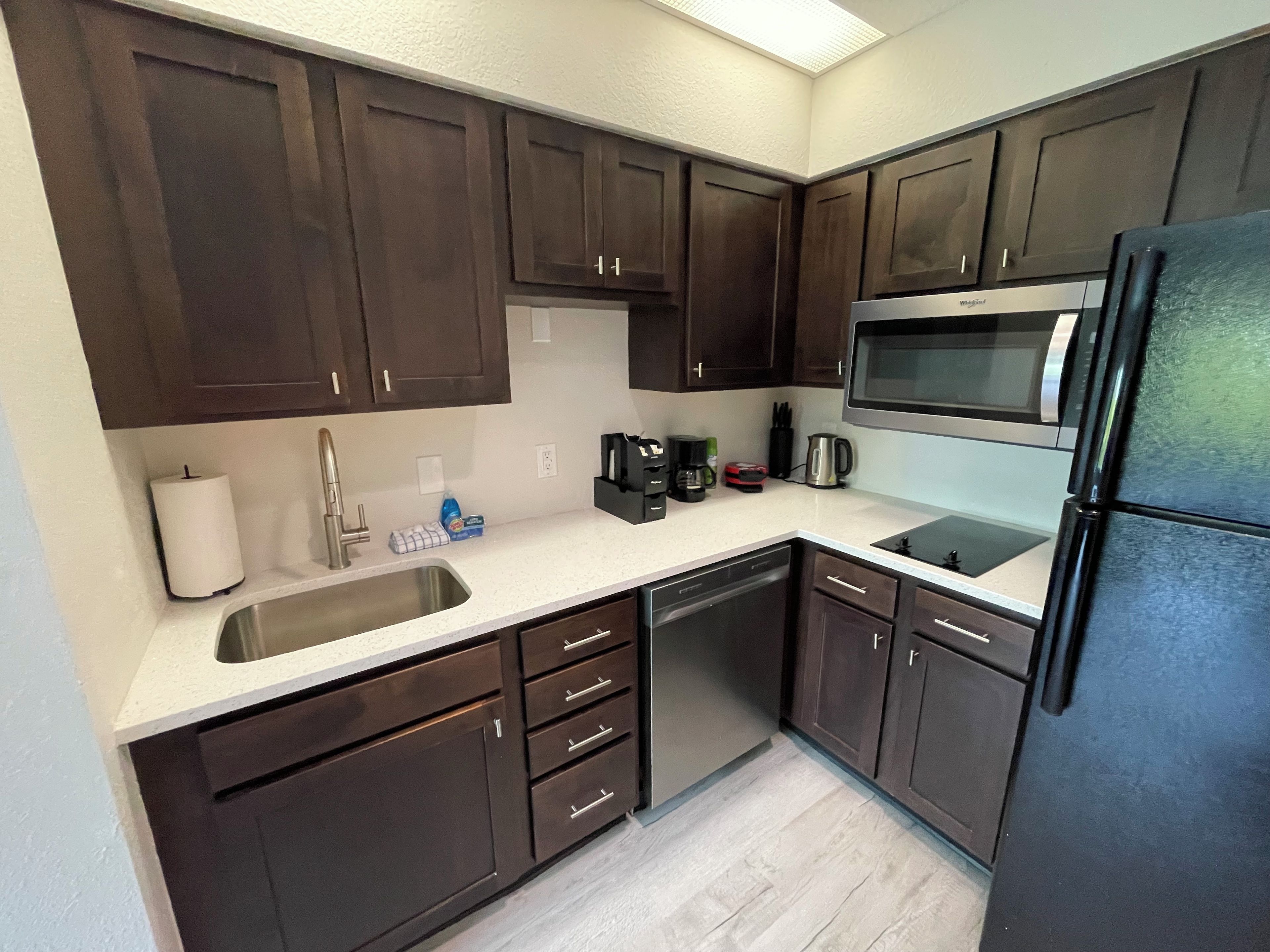 A compact kitchen with dark wooden cabinets, a stainless steel sink, a black refrigerator, and a microwave.