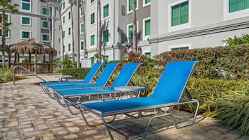 A poolside area with lounge chairs, a thatched structure, and a building with multiple windows and balconies.
