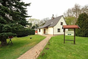 A two-story house with a paved driveway leading to the entrance, surrounded by a well-maintained lawn and a large tree. - Holiday home with 1 bedroom (Putbus\/ OT Kasnevitz)