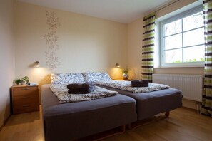 Een tweepersoonsbed met een gestreept hoofdeinde, bedlampjes, een klein nachtkastje met een plant en een raam met gordijnen.