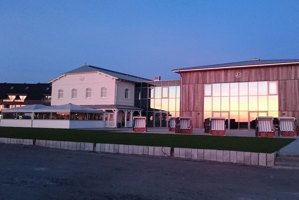 A building with a large glass facade, a covered outdoor seating area with chairs, and a grassy foreground.