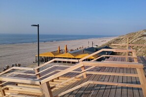 Ein hölzerner Steg führt zu Strandsonnenschirmen und einem Gebäude mit einem geneigten Dach.