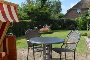 A patio with a round table and two chairs, a thatched-roof house in the background, and a hedge with pink flowers. - Holiday home in Nieblum with swimming pool (Nieblum)