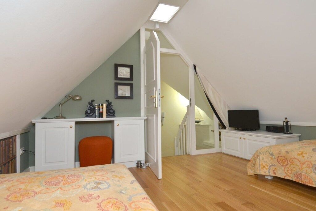 A bedroom with a sloped ceiling, a desk with a chair, a bed with a patterned bedspread, and a television on a stand.