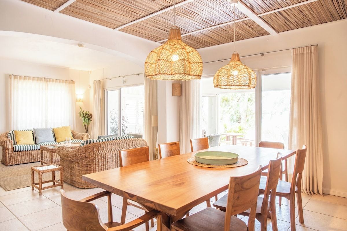 Un espace repas avec une table et des chaises en bois, un luminaire suspendu en fibres tressées et une vue sur l’extérieur grâce à de grandes fenêtres.