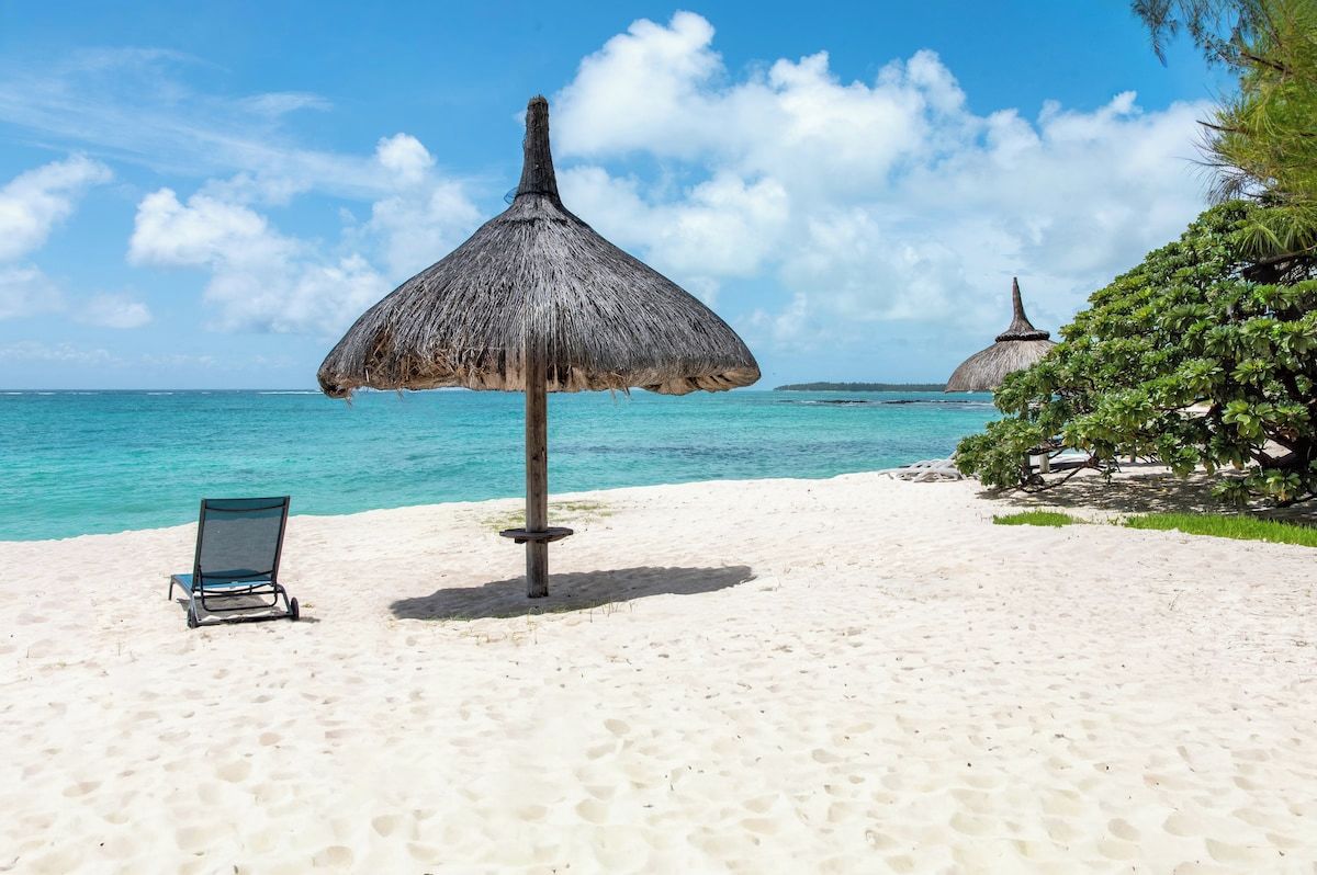 A beach with a thatched umbrella, a lounge chair, and a distant thatched structure.