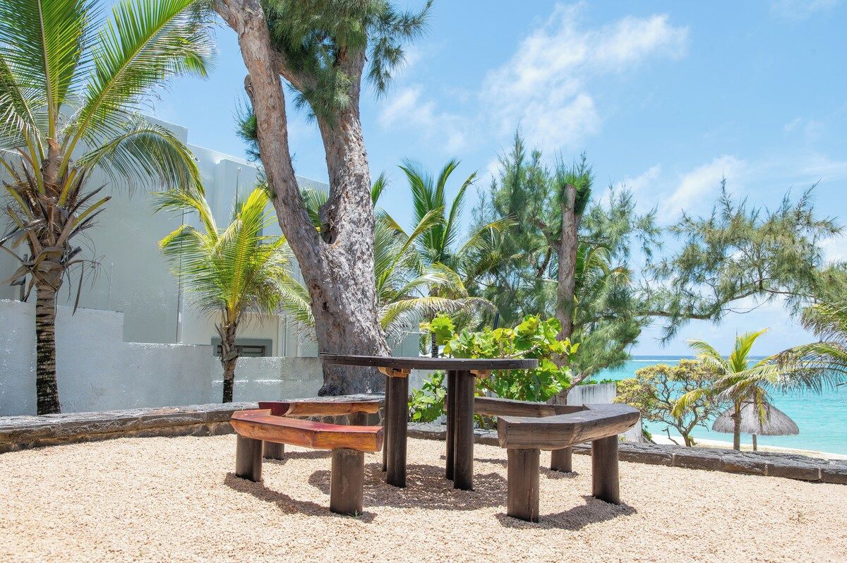 A beachside seating area with a table and benches, surrounded by palm trees and a view of the ocean.