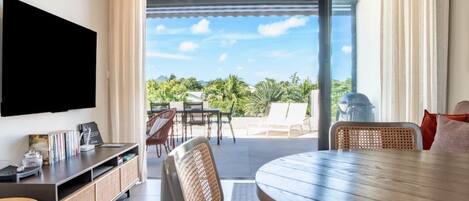 A modern hotel room with a flat-screen TV, a dining area with a round table, and a balcony with outdoor furniture.