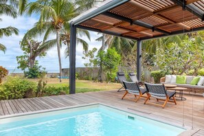 Un espace piscine comprenant des chaises longues, une terrasse en bois et un coin salon couvert avec des coussins.
