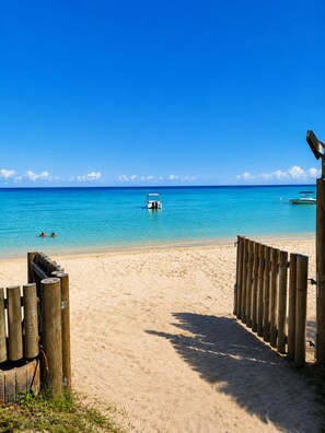 Une plage avec une clôture en bois, une eau d’un bleu limpide et un bateau à l’horizon.