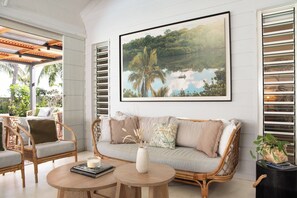 Une pièce comprenant un canapé en rotin, des chaises en osier, une table ronde en bois et un grand tableau encadré représentant un paysage tropical.