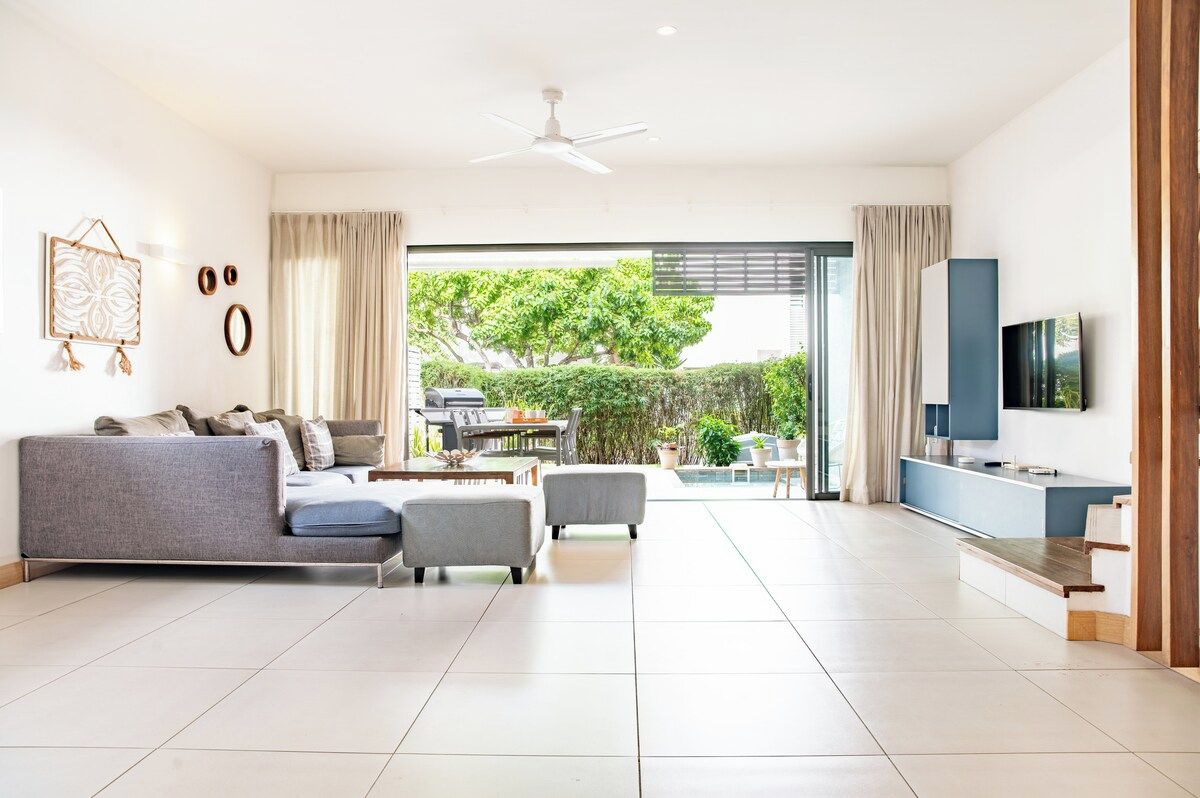 A modern living room with a sofa, armchairs, a TV, and a balcony with a view of greenery.
