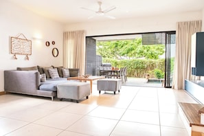 A modern living room with a sofa, ottoman, and coffee table, a flat-screen TV, and a sliding glass door leading to a patio with a grill. - Amazing Sunsets & Plunge Pool - Walk to Everything (Grande Riviere Noire)