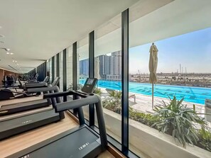 A gym with treadmills and exercise equipment, large windows, and a view of a swimming pool and buildings. - Trillionaire 1BR Trendy Timeless Tempting (Dubai)