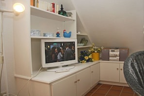 A room with a television on a white cabinet, a bookshelf with various items, and a decorative vase with flowers. - House on the Wadden Sea Holiday Apartment 8 (Sylt- Ost)