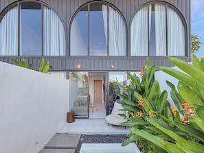 Een modern huis met grote, gebogen ramen, een zichtbaar interieur met een bank en een bijzettafeltje, en een tuin met tropische planten.