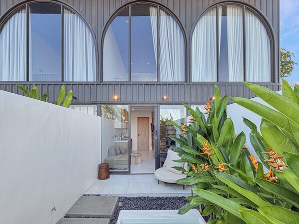 Ein modernes Haus mit großen Bogenfenstern, ein sichtbarer Innenraum mit einer Couch und einem kleinen Tisch sowie ein Gartenbereich mit tropischen Pflanzen.