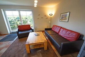 A living room with a sofa, a coffee table, and a floor lamp. There is a window with a view of greenery outside. - Holiday home in Prerow with barbecue (Prerow)