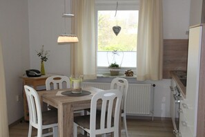 A dining area with a wooden table and white chairs, a hanging light fixture, a window with curtains, and a radiator. - Holiday apartment in Büsum with swimming pool (Büsum)