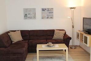 A living room with a brown sofa, a small wooden coffee table, a floor lamp, and wall art. - Holiday apartment in Büsum with swimming pool (Büsum)