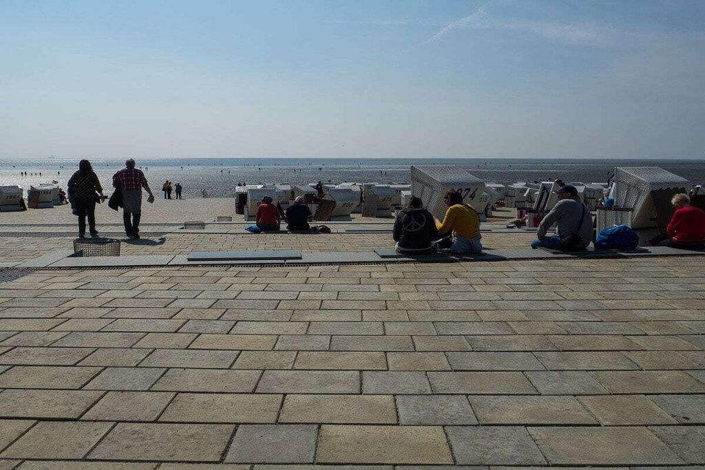 Un vialetto lastricato che conduce a una spiaggia, con persone sedute e in piedi, e ombrelloni da spiaggia in lontananza.