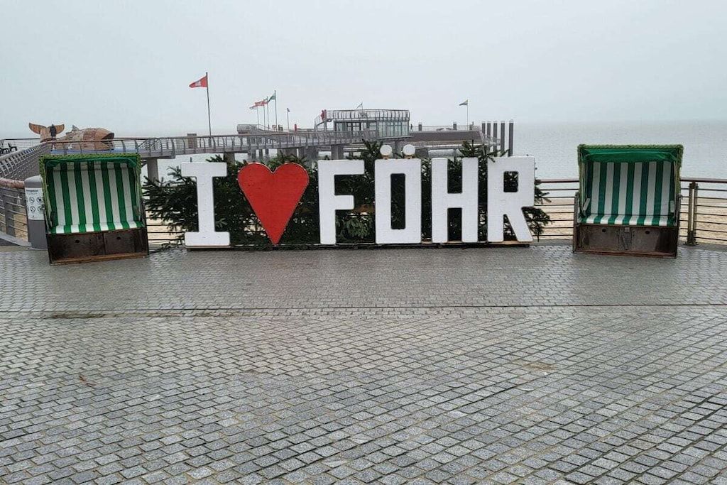 A sign with 'I ♥ FOHR' in large letters, two green-striped chairs, and a view of the sea.
