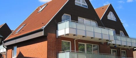 Un immeuble résidentiel de deux étages avec une façade en briques, un balcon doté de rampes en verre et un toit recouvert de tuiles rouges.