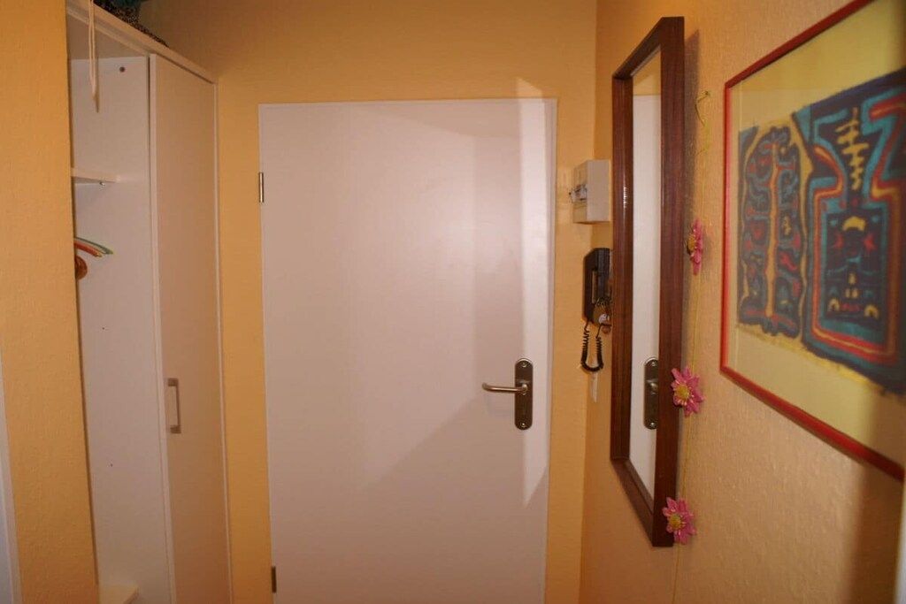 Eine geschlossene Tür mit einem schlichten Griff, ein weißer Kleiderschrank links und ein gerahmtes Kunstwerk mit floralem Rand an der Wand.