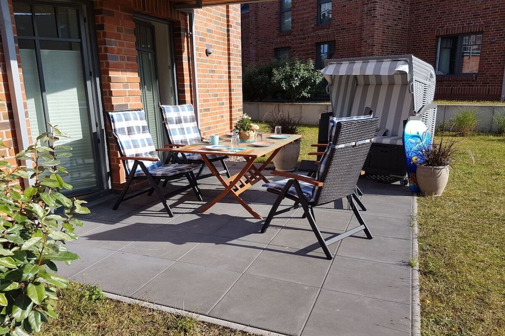 Une terrasse avec une table et des chaises, un barbecue abrité et, au fond, un bâtiment en briques.
