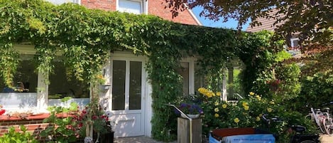 A residential building with a brick facade, a white door, and a garden with various plants and flowers.