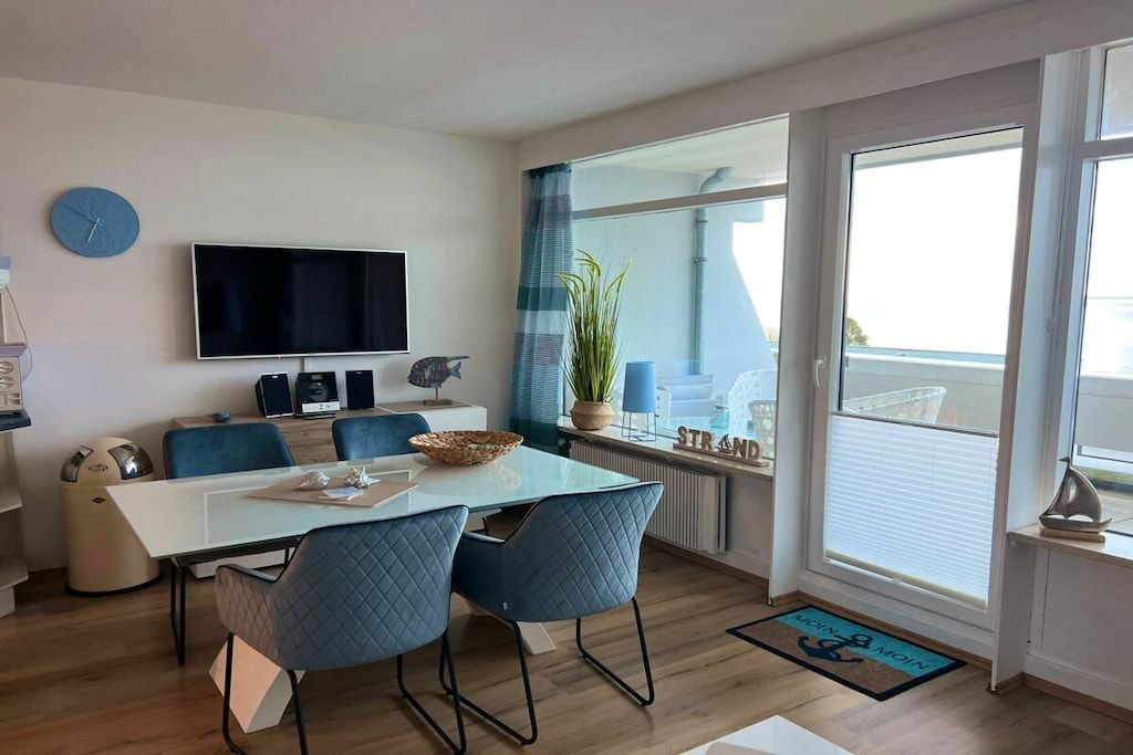 A modern living room with a flat-screen TV, a dining table with chairs, a large window with curtains, and a view of the outside.