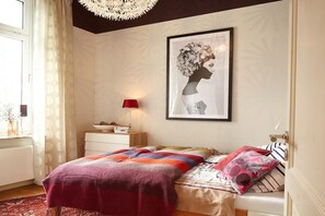 Une chambre à coucher avec un lit, une table de chevet, une lampe, une œuvre d’art encadrée au mur et une fenêtre avec des rideaux.
