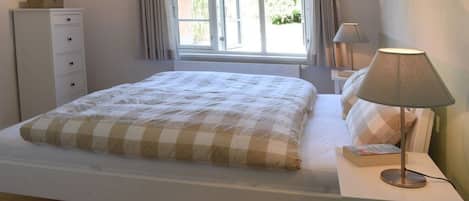 Ein Schlafzimmer mit einem Bett, einem Nachttisch mit Lampe, einer Kommode und einem Fenster mit Vorhängen.