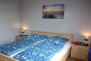 A double bed with a blue patterned bedspread, two bedside tables with lamps, and a painting of a sunset on the wall. - "Sun-filled apartment on the Schlei" (Akeby)